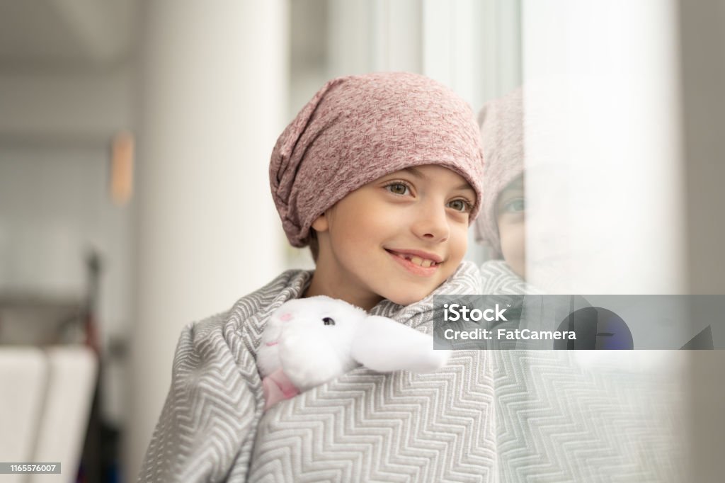 Smiling girl with cancer looks out window A happy elementary age girl with cancer is wearing a pink scarf on her head. She has a blanket draped over her shoulders and is holding a stuffed toy. The child is smiling boldly while leaning against a window. Child Stock Photo