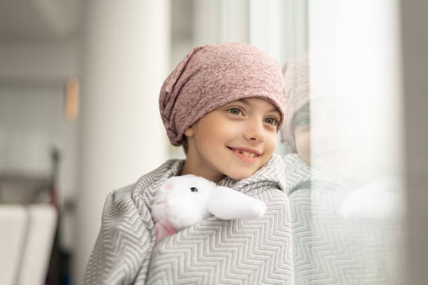 Smiling girl with cancer looks out window A happy elementary age girl with cancer is wearing a pink scarf on her head. She has a blanket draped over her shoulders and is holding a stuffed toy. The child is smiling boldly while leaning against a window. cancer illness stock pictures, royalty-free photos & images