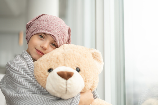 An cute elementary age girl with cancer sits by a window. She has cancer and is wearing a pink bandana. The child is wrapped in a blanket and is hugging a large teddy bear. She is smiling at the camera.