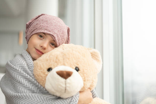 Portrait of girl with cancer holding a teddy bear An cute elementary age girl with cancer sits by a window. She has cancer and is wearing a pink bandana. The child is wrapped in a blanket and is hugging a large teddy bear. She is smiling at the camera. cancer illness stock pictures, royalty-free photos & images