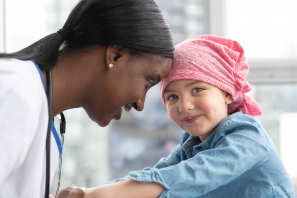 Female doctor comforts her young patient who has cancer A cute elementary age girl with cancer is wearing a pink scarf on her head. She is at a medical appointment. The female doctor of African descent is holding the child's hands, providing comfort and support. The child is smiling at the camera. paediatrician stock pictures, royalty-free photos & images