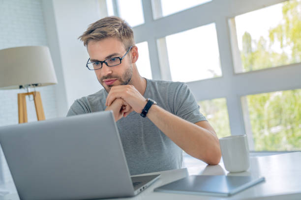 istock Concentrated serious handsome man in eyeglasses using laptop computer while sitting by the table at office 1165582870