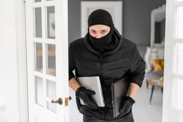 thief looking at the camera with black balaclava stealing two modern expensive laptops. the burglar commits a crime in luxury apartment with stucco. - night deposit box imagens e fotografias de stock
