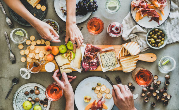 Summer picnic with rose wine and snacks and peoples hands Mid-summer picnic with wine and snacks. Flat-lay of charcuterie and cheese board, rose wine, nuts, olives and peoples hands over concrete table background, top view. Family, friends holiday gathering salumeria stock pictures, royalty-free photos & images