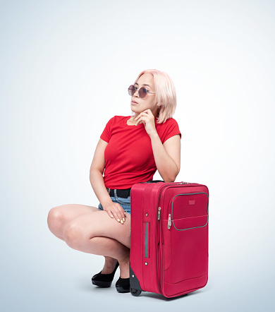 Young blonde girl in sunglasses and a red T-shirt is sitting next to a suitcase waiting for a flight, on light background.