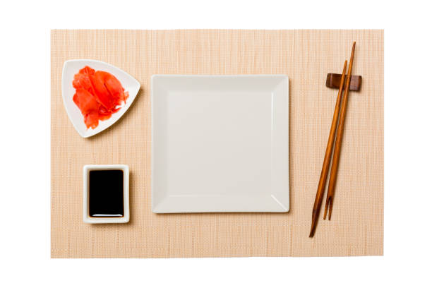 Empty white square plate with chopsticks for sushi and soy sauce, ginger on brown sushi mat background. Top view with copy space for you design.