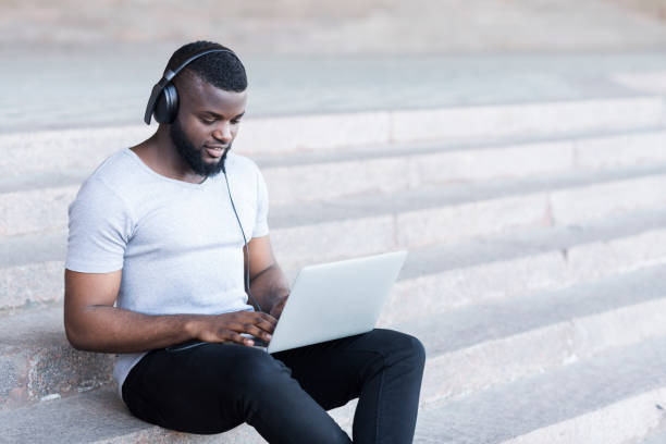 istock Young bearded african guy in headphones coding on laptop 1165681689