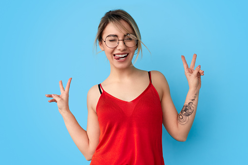 Excited young woman in trendy outfit and glasses winking and showing tongue while gesturing V sign against blue background