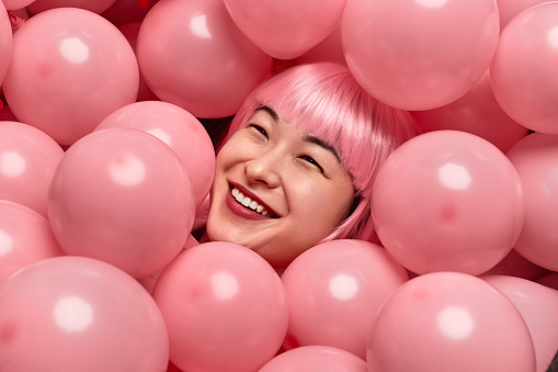 From above excited Chinese female in pink wig cheerfully smiling while lying under heap of balloons and having fun