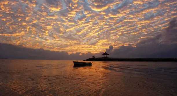 Fishing boat silhouette floating and the famous gazebo icon of Sanur Beach under beautiful dramatic sunrise sky. Clouds abstract pattern texture. Sunrise glow & nature beach landscape background.