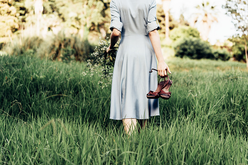 Rear view of a woman in a dress walking along a meadow barefoot with shoes in her hand.