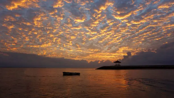 Fishing boat silhouette floating and the famous gazebo icon of Sanur Beach under beautiful dramatic sunrise sky. Sunrise glow & nature beach landscape background.