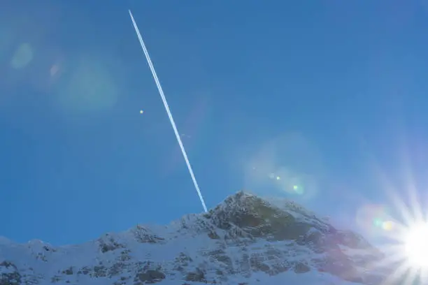 An aircraft with its contrail raising behind a big rocky mountain and the sun with its star-shaped rays as backlight