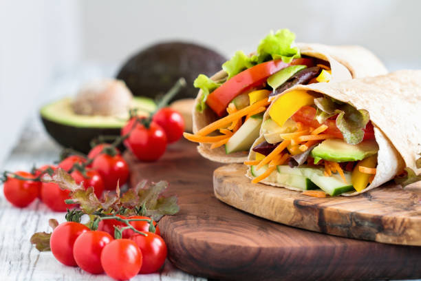 Healthy Vegan / Vegetarian Veggie Wrap A healthy lunch or dinner of a vegan / vegetarian wrap made with  argula lettuce, sliced tomatoes, cucumbers, avocado, bell peppers and carrots. Selective focus on sandwich in front. wrap sandwich photos stock pictures, royalty-free photos & images