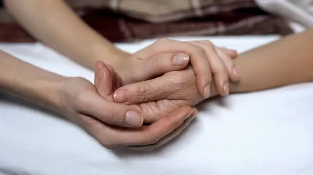 Photo of Warm daughters hands holding and calming down sick mother in bed, assistance