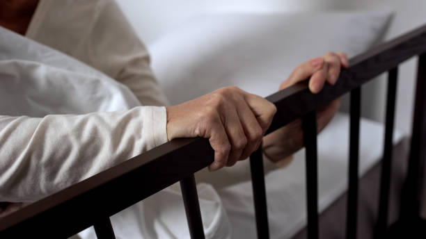 Aged ill woman hardly getting out of bed holding on railing, hospital ward stock photo