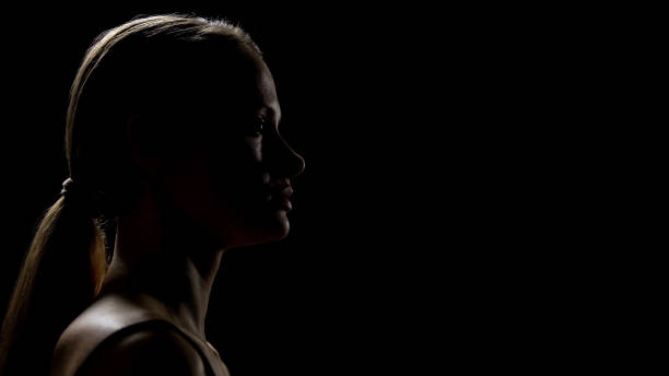 Portrait of a young woman with wavy blonde hair looking down, showing natural beauty, shiny healthy strands, soft skin texture and calm emotional mood on a dark background