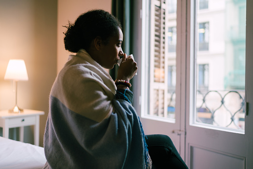 Young woman sitting at the bed with cup of tea and looking through the window