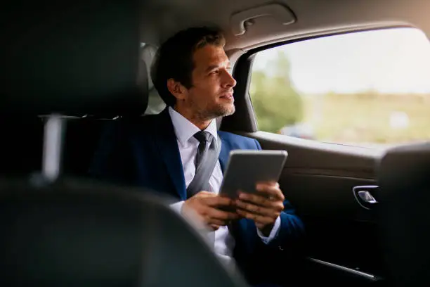 Handsome businessman sitting with digital tablet on the backseat of the car stock photo. Shadow DOF. Developed from RAW; retouched with special care and attention; Small amount of grain added for best final impression. 16 bit Adobe RGB color profile.