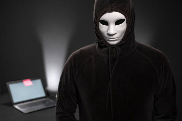 istock Hooded computer hacker with mask looking at the camera in front of a desk with laptop 1165712995