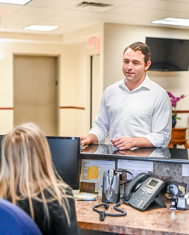 Handsome man in his 30’s stands at a office reception desk and speaks to a female receptionist. She is sitting at her desk and the man may be a client, customer, or a socializing co-worker. Office setting