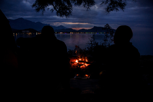 Silhouettes two people sitting on the seacoast near the campfire in the dark looking at the Kemer, Turkey on the other beach