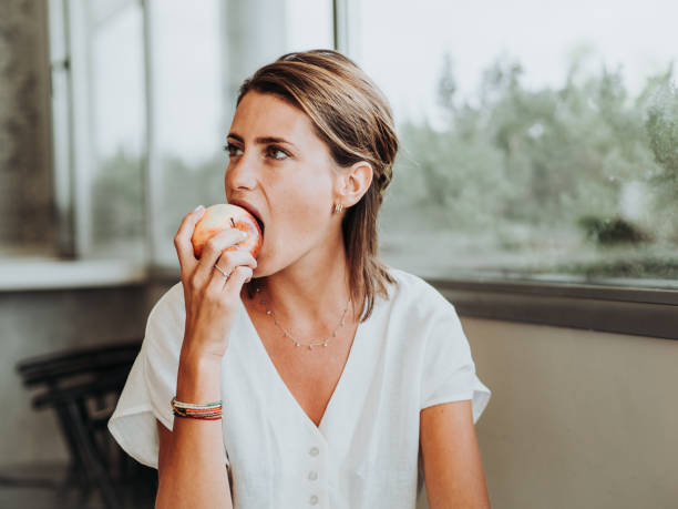 giovane donna che mangia una mela mordendo davanti alla grande finestra della cucina di casa sua - apple women green eating foto e immagini stock