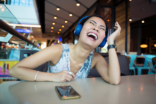Excited and surprised Asian woman listening to podcast by mobile phone, smiling in coffee shop