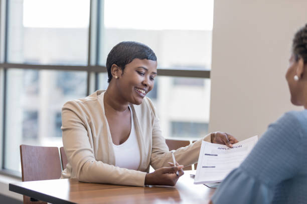 Personal banker explains account application to client A female personal banker sits across a desk from her client and gestures to a banking application as she gives instructions. bank teller stock pictures, royalty-free photos & images