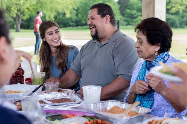 Cheerful man talks with relatives during family reunion Smiling mature Hispanic man laughs while talking with family members during a lunch in a park. pot luck stock pictures, royalty-free photos & images
