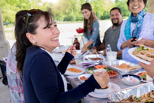 Hispanic woman laughs while talking with a relative during a family picnic in the park.