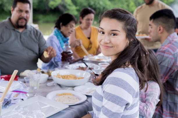 Beautiful young woman enjoying meal during family gathering Young Hispanic woman smiles while looking at the camera during an outdoor meal. big family table stock pictures, royalty-free photos & images