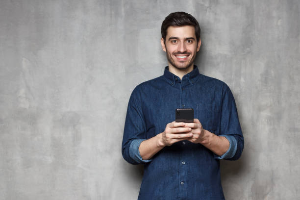 istock Smiiling modern business man  standing against gray textured wall, holding his phone with both hands. Copy space on the left side 1165763883