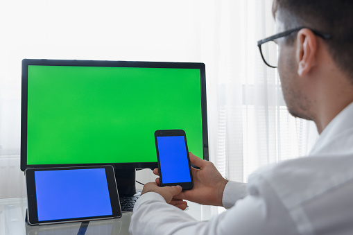 Engineer, Constructor, Designer in Glasses Working on a Personal Computer with a Green Screen on Monitor which has Chroma Key Great for Mockup Template. Male Using Smartphone or Tablet with Blue Screen and Creating, Designing using CAD Software. Freelance Work.