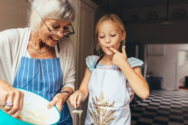Grandmother and kid having fun making cake in kitchen Senior woman in apron making batter for cake. Little girl tasting cake batter standing in kitchen with grandmother. granddaughter stock pictures, royalty-free photos & images