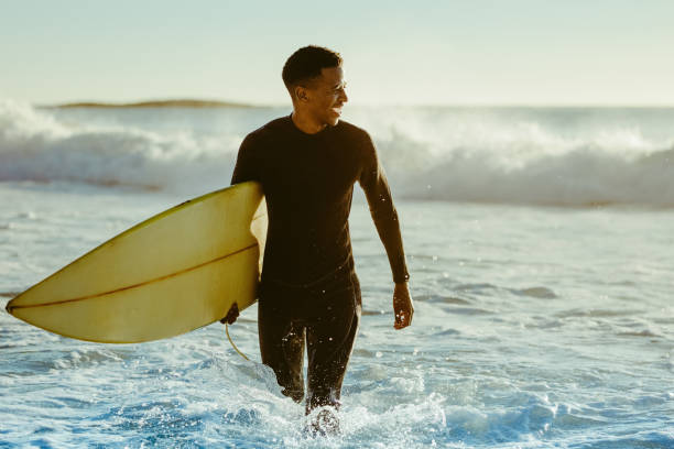 Surfer coming out of the ocean Smiling young man with surfboard on the beach. African male coming out of the ocean after water surfing. surfing stock pictures, royalty-free photos & images