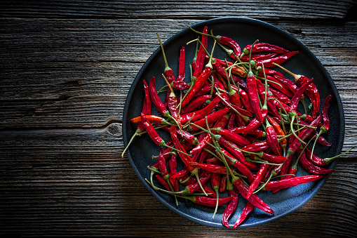 Dried organic red chili peppers in a black plate shot from above on rustic wooden table. The composition is at the right of an horizontal frame leaving a useful copy space for text and/or logo at the left. Low key 42Mp studio photo taken with SONY A7rII and Sony FE 90mm f2.8 Macro G OSS lens