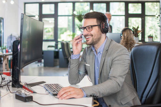 operatore di assistenza clienti sorridente che indossa cuffie che parlano con il cliente mentre lavora sul computer in ufficio - sales agent foto e immagini stock