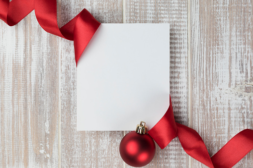 White blank card on white wooden background wrapped with a red ribbon. At the right bottom corner of the blank card is one red Christmas ball.