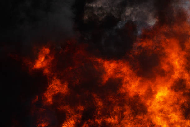 Dangerous red flames of strong industrial fire and dramatic black clouds of smoke covered sky. Atmospheric dispersion, soft and selective focus, motion blur from fire, high temperature from flames.