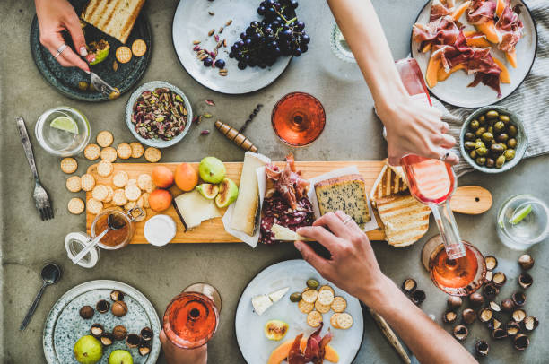 Summer picnic with wine and snacks and peoples hands Mid-summer picnic with wine and snacks. Flat-lay of charcuterie and cheese board, rose wine, nuts, olives and peoples hands holding food and celebrating over concrete table background, top view salumeria stock pictures, royalty-free photos & images