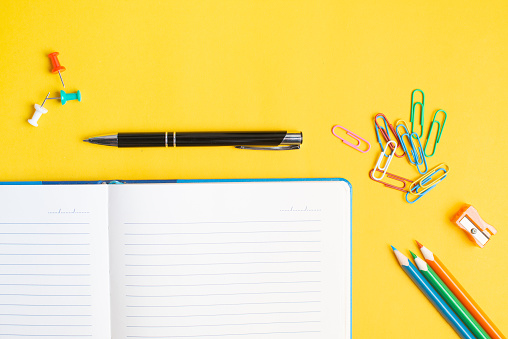 Notepad with pen and colorful pencils on yellow background. Colorful paper clips. Back to school concept. Copyspace.