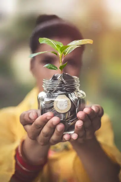 Close up of Indian female hands holding money coins in glass jar with plant in her hand for financial and saving
