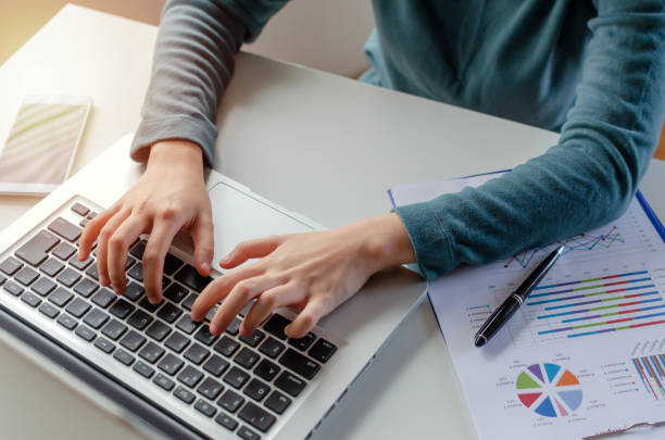 young woman typing on keyboard working and using laptop and graph statistics document on desk at home office, searching web, social network, online, finance, investment and digital technology concept