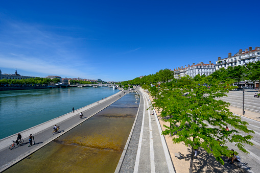 People walking, running and using bicycle along the Rhone river in central Lyon, France, a sunny summer day. Next to the river residential buildings are showing.