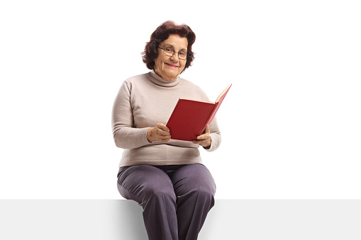 Senior woman with holding an open book and sitting on a panel isolated on white background