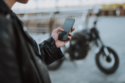 Man in leather jacket using modern smartphone. Motorbike on the background