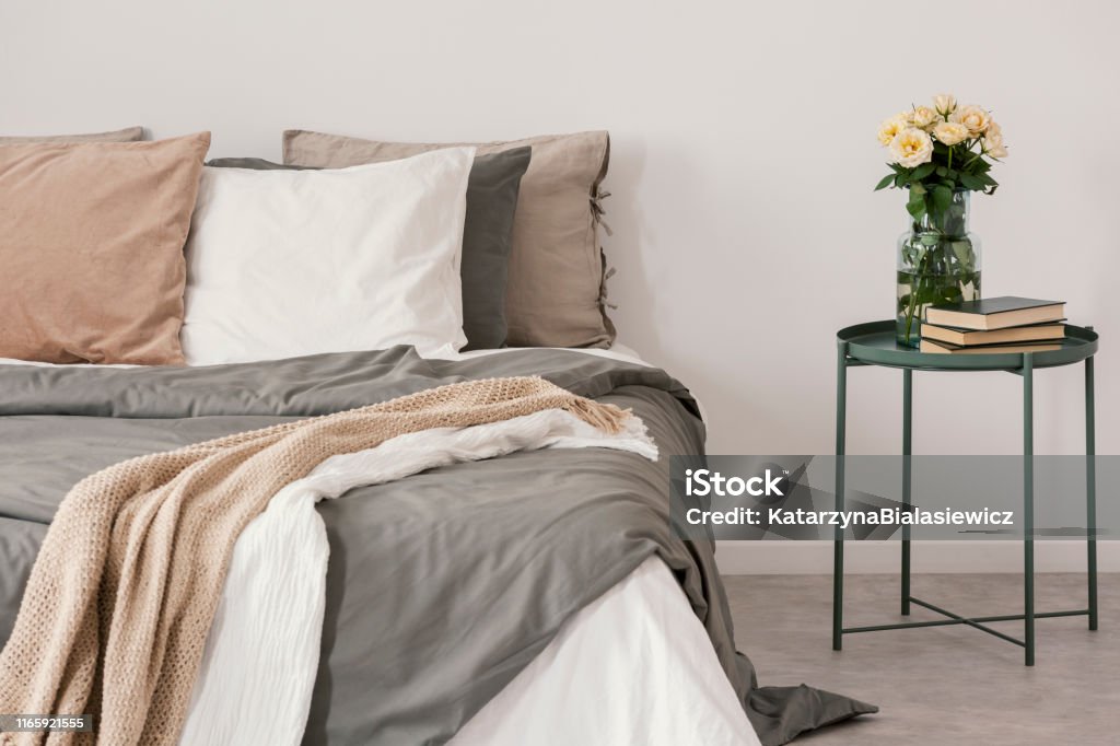 White roses in glass vase and books on stylish nightstand next to big bed with linen pillows and cozy blanket Linen Stock Photo