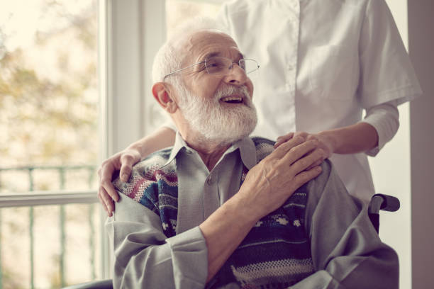 hombre mayor sentado en la silla de ruedas, riendo y sosteniendo la mano de su enfermera - asistente sanitario fotos fotografías e imágenes de stock