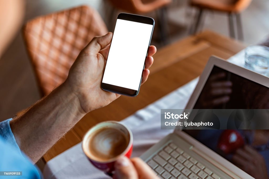 Getting the most out of the work day with technology Close-up. Businessman drinking a coffee and looking in phone in restaurant. Photo of a man using blank laptop sending massages on the coffee shop. Concept of success and well-being. Close up selective focus on smartphone in hands of young businessman sitting in cafe during the day. Entrepreneur working on business project in city cafe, using mobile phone Device Screen Stock Photo